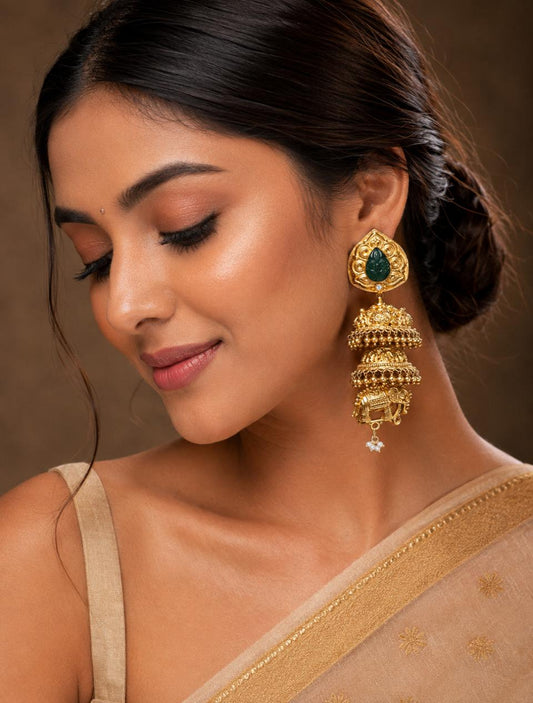 A close-up of a woman wearing an ornate gold jhumka earring with a green stone and a small white dangling accent, paired with a beige saree against a warm brown background.